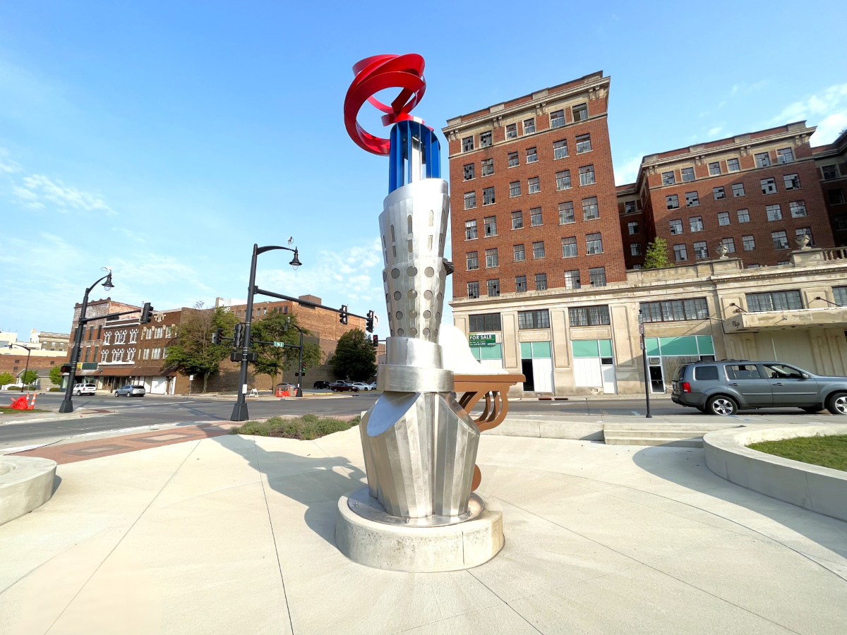 A public art piece representing a stylized torch with flame on top, sitting in the center of a concrete circle with old city buildings in the background