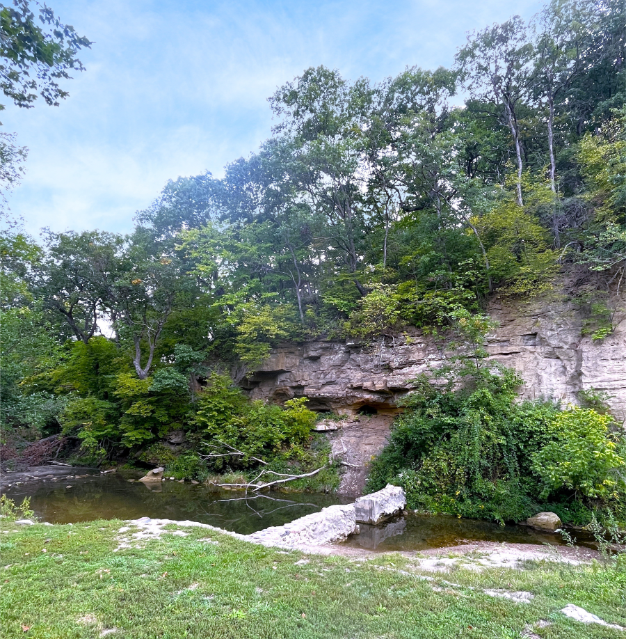 A landscape of forested cliff face with a creek at its base, bright blue sky, and manicured grass in the foreground. Dolliver park.