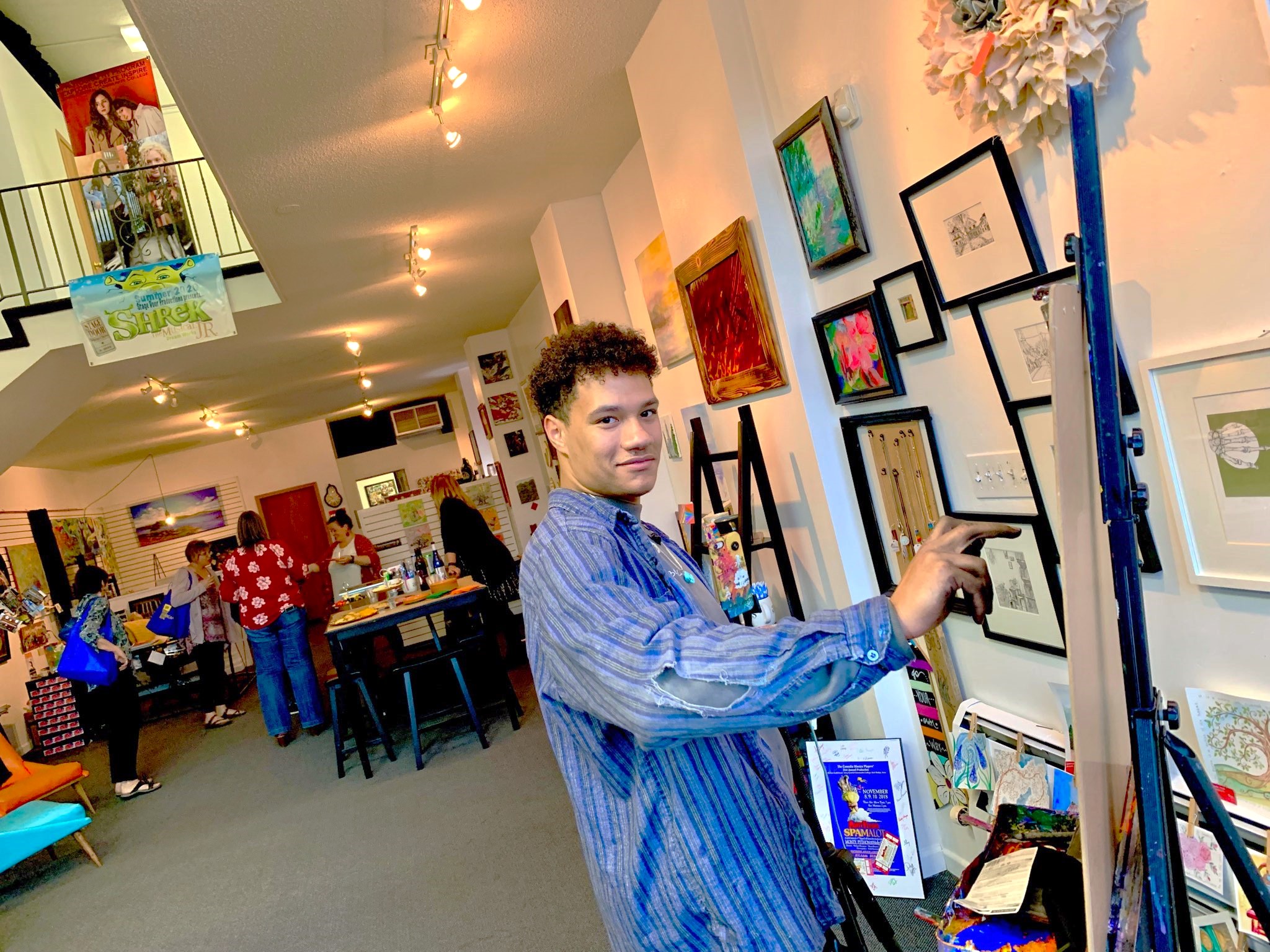A young, male artist stands at his easel doing charcoal art. Fine art hangs on the walls around him and a group of ladies in the background chat while crafting.