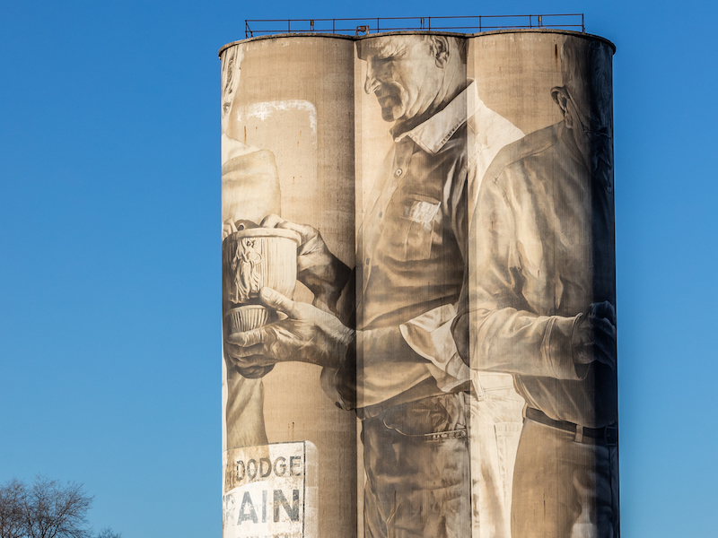 Three conjoined silos bearing a full-length painted mural of a man holding pottery stand out against a bright blue sky.