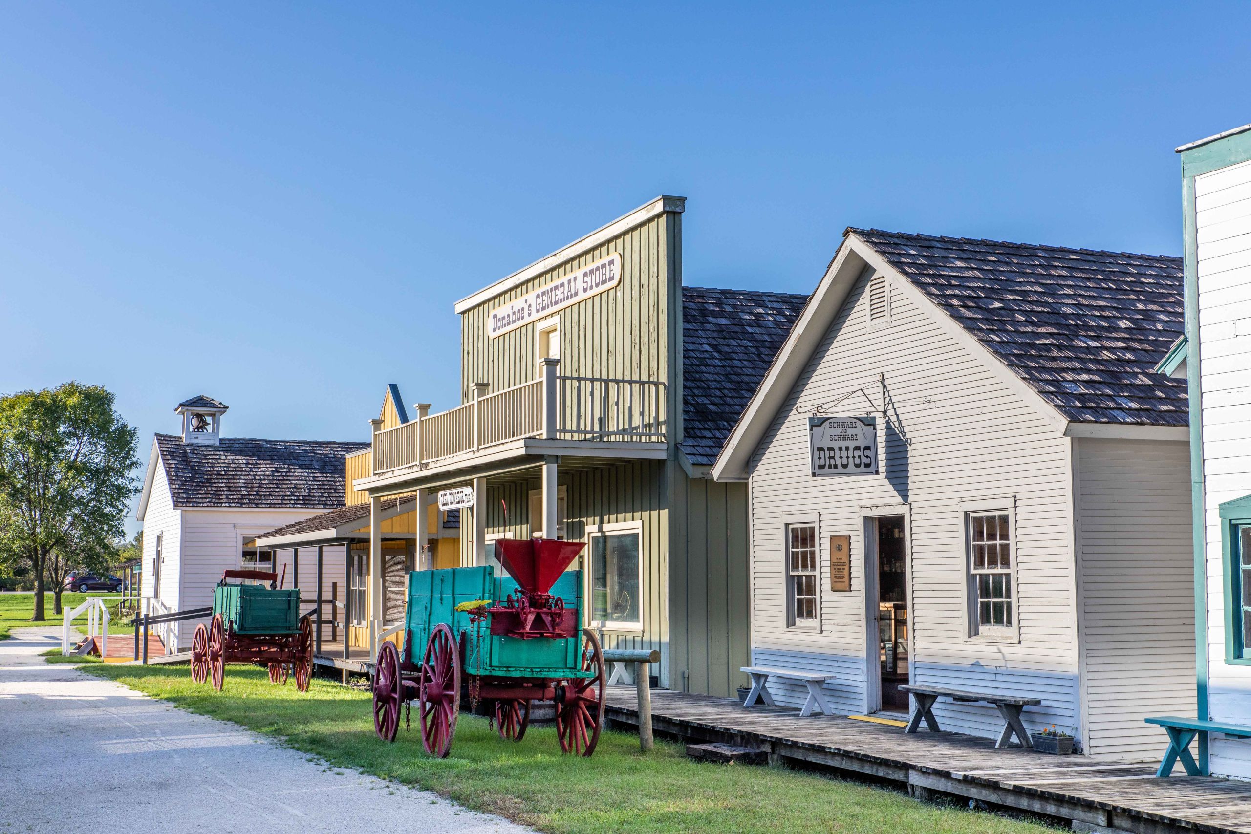 Several old-timey buildings are lit by morning sun with a blue sky and clean gravel road in the foreground. Fort Museum.