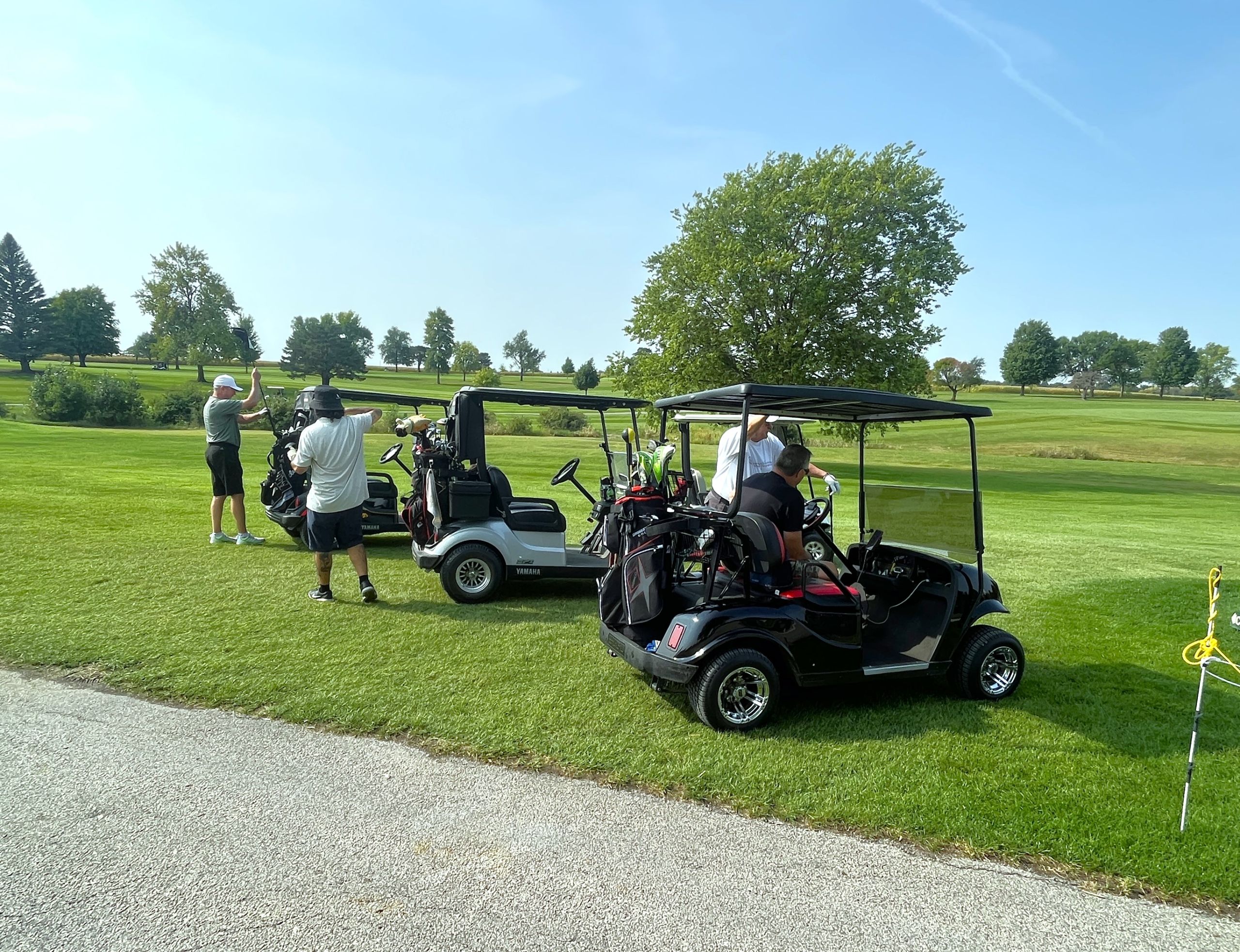 Three golf carts are lined up on a lakeside green with men preparing to play golf.