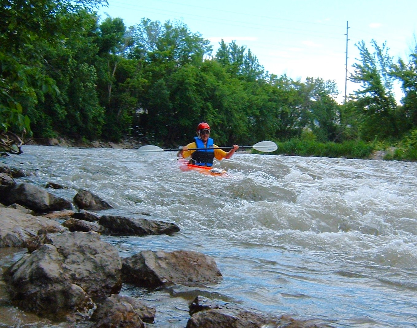 A man in an orange kayak and blue life vest navigates rapids near the shore of a water trail with forest in the background.