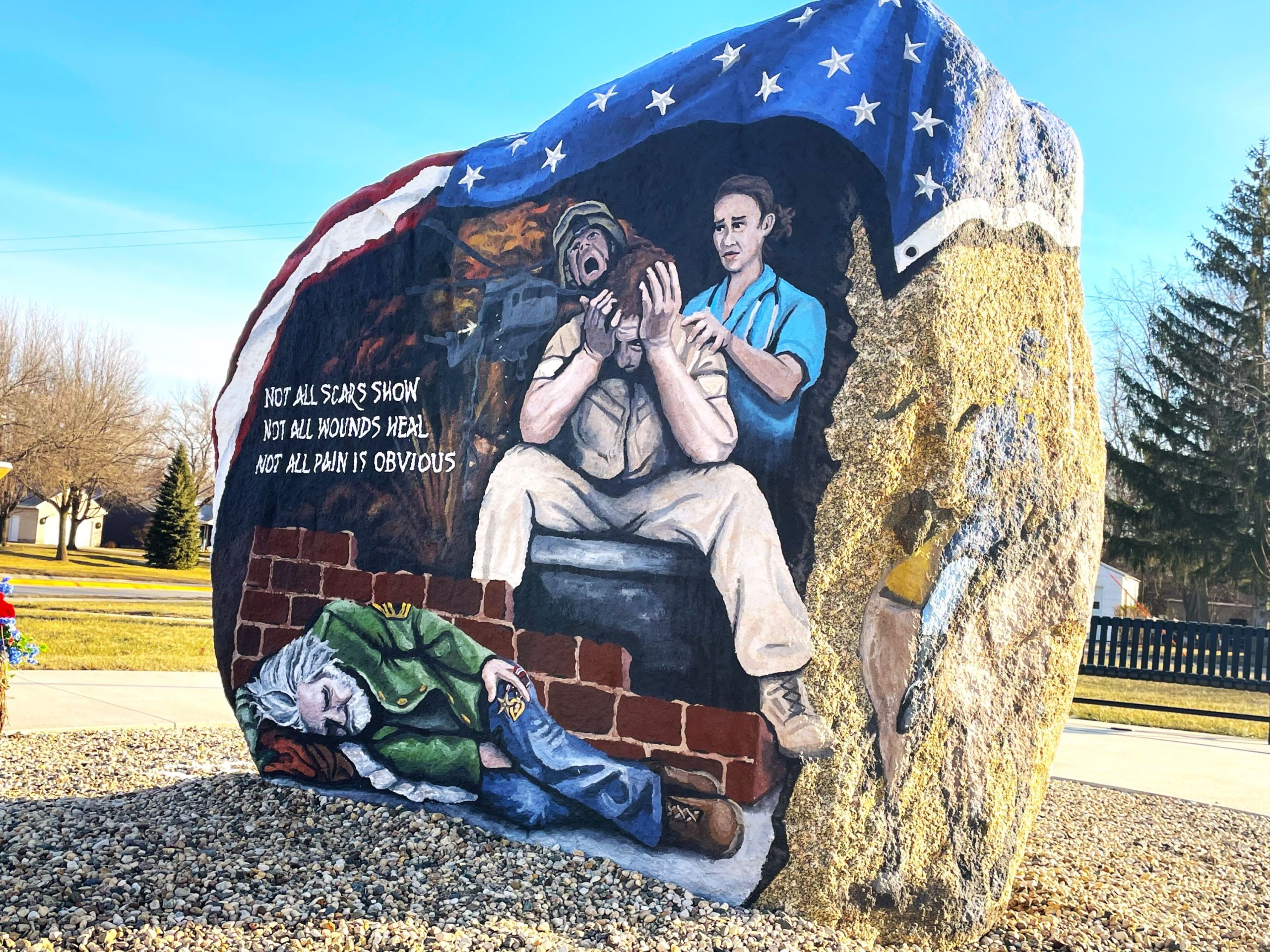 A large boulder of granite is painted with patriotic scenes, featuring a veteran experiencing PTSD and a homeless man in uniform.