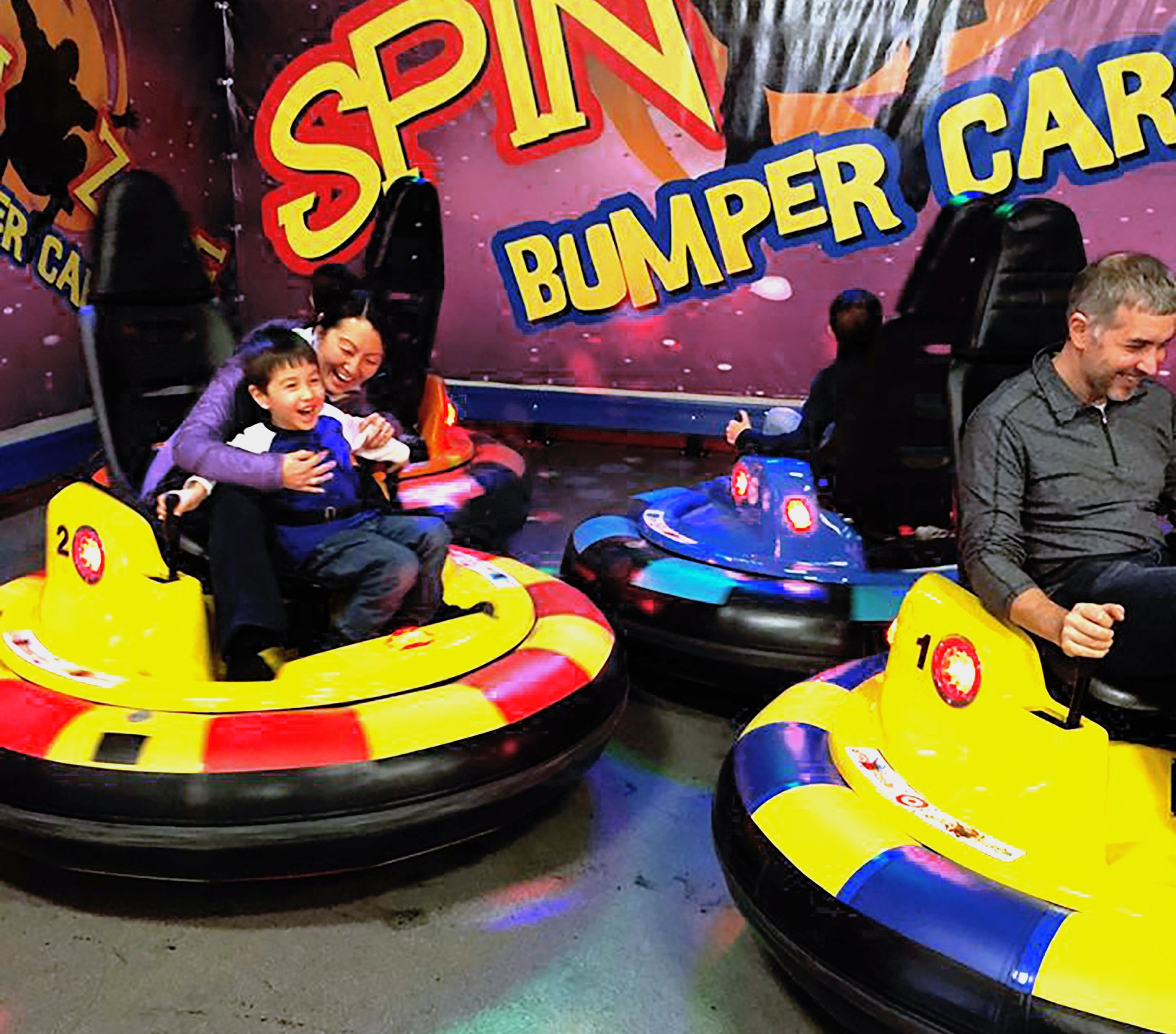 A mother and son ride a bumper car, both smiling and having family fun. They are about to bump into a man on another car.