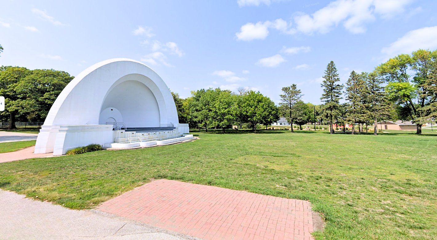 A white acoustic bandshell sits on a manicured green space with a brick patio in the foreground, trees and other buildings in the background. Oleson Park.