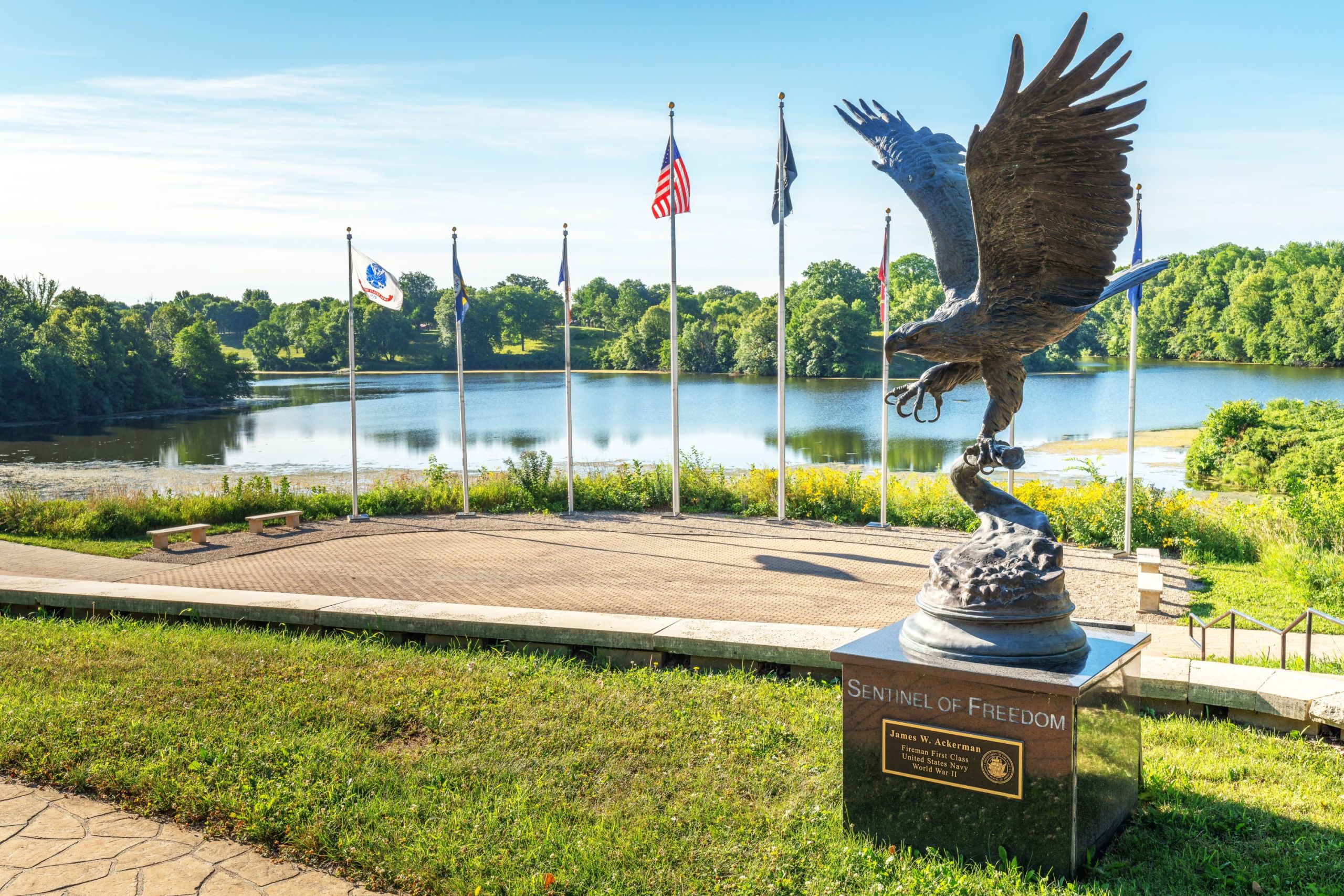 A bronze statue of an eagle with wings raised overlooks the outdoor amphitheater of a park flanked by military and national flags. This sits by a lake with forest on the far shore.