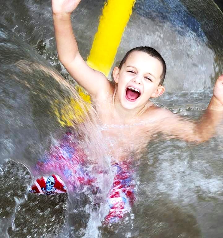 A young boy in swim trunks is bombarded with water spouts from a splash pad and laughs.