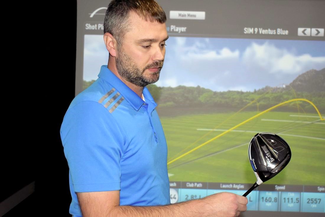 man in blue shirt standing in front of golf simulator screen, inspecting a black golf club