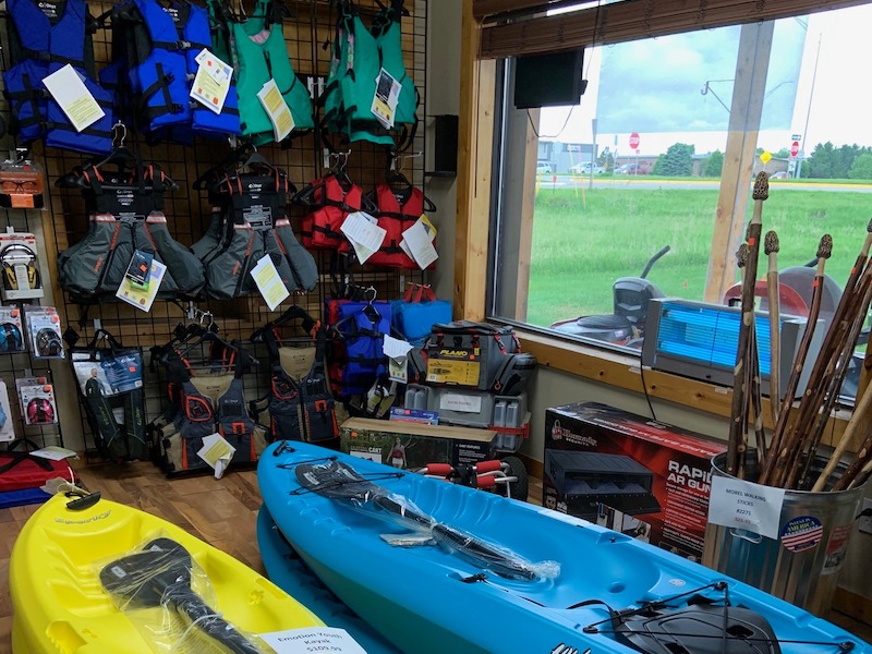 Wall with blue, green, grey, and red life vests, yellow and blue kayaks on floor, other assorted outdoors recreation equipment.