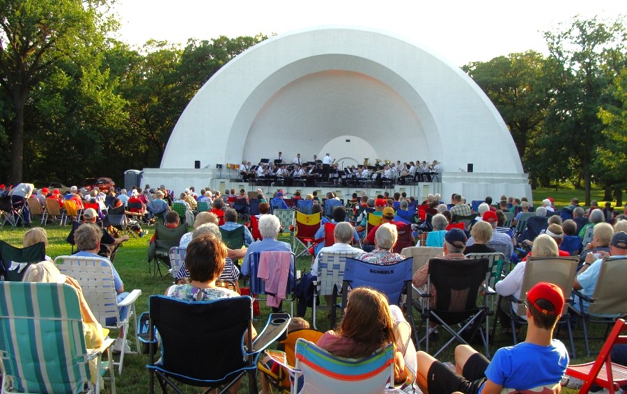 A crowd of people sit in camp chairs and on blankets watching a municipal band play in an acoustic bandshell, surrounded by trees. Oleson Bandshell.