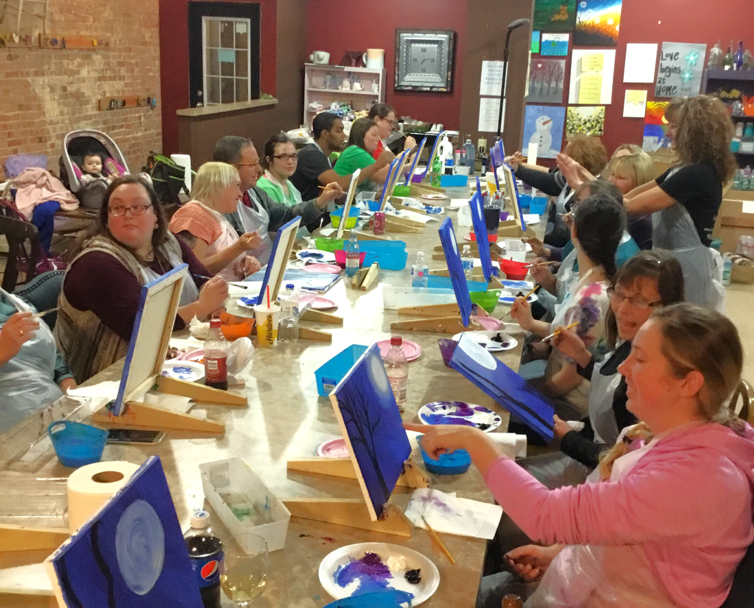 A group of men, women, and children sit at a long table painting on canvases. They are surrounded by art supplies and completed works on the walls of the studio.